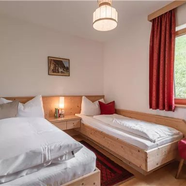 Ein gemütliches Schlafzimmer mit zwei Einzelbetten, Holzkommode und einem Stuhl. Große Fenster lassen viel Tageslicht herein, und rote Vorhänge verleihen dem Raum eine warme Atmosphäre.