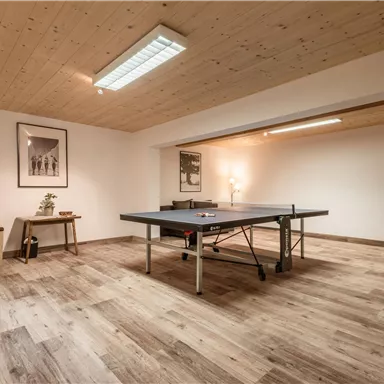 Ein heller Raum mit einem Tischtennistisch in der Mitte und einem bequemen Sessel in der Ecke. Die Wände sind einfach gestaltet und der Boden ist aus Holz.