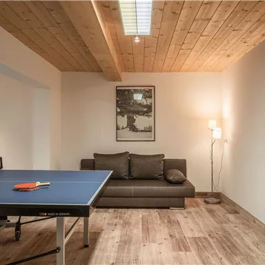 Ein gemütlicher Raum mit einem Tischtennistisch und einem Sofa. Die Wände sind minimalistisch dekoriert, und es gibt eine Stehlampe für zusätzliche Beleuchtung.