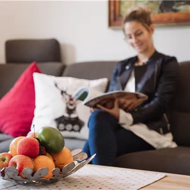 Eine gemütliche Wohnsitz mit einer sitzenden Frau, die ein Magazin liest. Auf dem Tisch steht eine Schale mit buntem Obst.