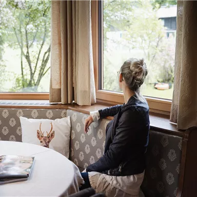 Eine Frau sitzt an einem Tisch und schaut aus dem Fenster. Auf dem Tisch stehen Rosen und ein offenes Buch ist sichtbar.