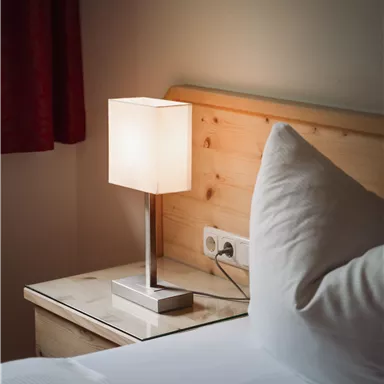Eine moderne Nachttischlampe steht auf einem Tisch neben einem Bett. Im Hintergrund ist ein Holzpaneel und ein gemütliches Kopfkissen zu sehen.