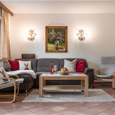 Ein gemütliches Wohnzimmer mit einer Couch und zwei Sesseln. An der Wand hängt ein Gemälde, und ein Tisch ist mit Früchten dekoriert.