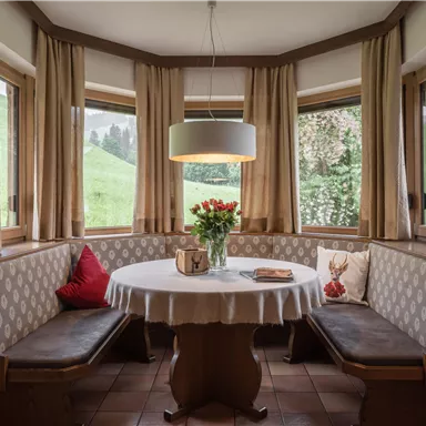 Ein gemütlicher Essbereich mit einem runden Tisch und Bänken. Große Fenster bieten einen Blick ins Grüne und frische Blumen schmücken den Tisch.