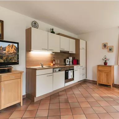 Eine helle Küche mit modernen Schränken und einem großen Fernseher. Der Raum hat Fliesenboden und ist freundlich eingerichtet.