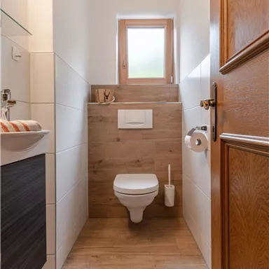 Ein modernes Badezimmer mit WC, Waschbecken und Holzdetails. Es hat viel Licht durch ein Fenster und eine helle, einladende Atmosphäre.