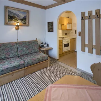 Ein gemütlicher Wohnraum mit einem blumigen Sofa und einem Esstisch. Die Küche ist durch einen Bogen sichtbar und es gibt Holzpaneele an der Wand.