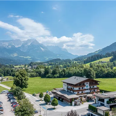 Ein malerisches Bergland mit grünen Wiesen und majestätischen Bergen im Hintergrund. Im Vordergrund ist ein traditionelles Chalet zu sehen, umgeben von Bäumen und Parkplatz.