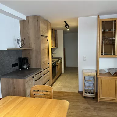 Eine moderne Küche mit Holzregalen und einem Esstisch. Der Raum ist hell und ordentlich, mit Geräten und Geschirr im Hintergrund.