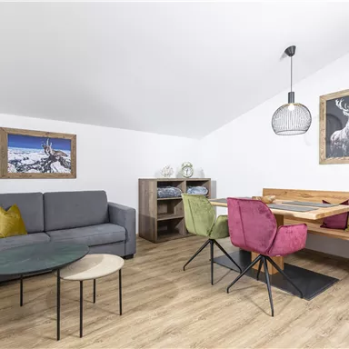 Ein modernes Wohnzimmer mit einem grauen Sofa und bunten Kissen. Ein Esstisch mit Stühlen und dekorativen Bildern an der Wand.