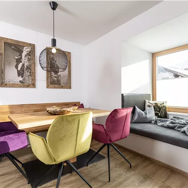 Ein moderner Essbereich mit einem holzfarbenen Tisch und bunten Stühlen. Große Fenster lassen viel Licht herein und bieten einen Blick nach draußen.