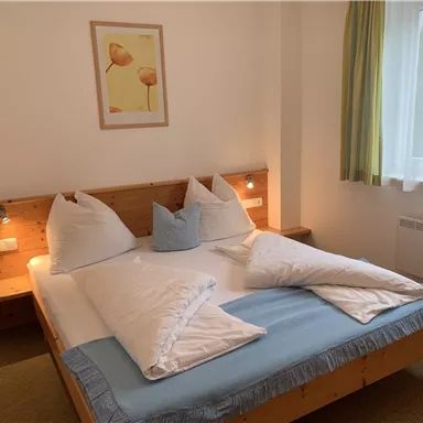 Ein gemütliches Schlafzimmer mit einem Holzrahmenbett und zwei großen Kissen. Das Zimmer hat ein freundliches Ambiente mit einem Fenster und einem bunten Bild an der Wand.