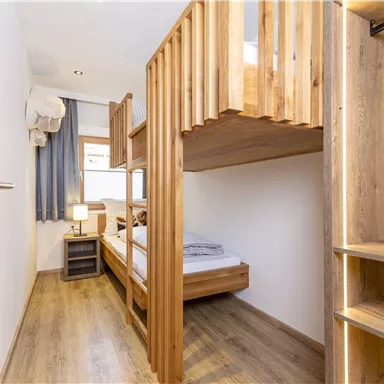 Ein funktionales Zimmer mit Holzbetten und einem kleinen Nachttisch. Viel Tageslicht kommt durch das Fenster, und der Boden ist mit Holzplatten ausgelegt.