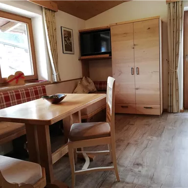 A cozy living space with a wooden table, chairs, and a bench. Large windows let in plenty of light and there is a cabinet with a television.