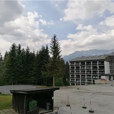 A tranquil landscape with trees and a modern building. In the background, mountains and a cloudy sky are visible.