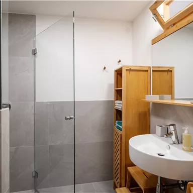 A modern bathroom with a tiled shower and a sink. Wooden shelves provide space for towels and toiletries.