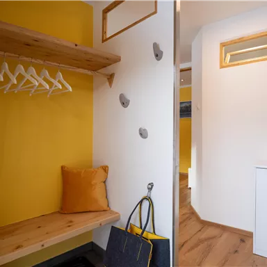 A bright entrance area with a wooden bench and yellow walls. Coat hooks are hanging on the wall, and there is a small chest of drawers.