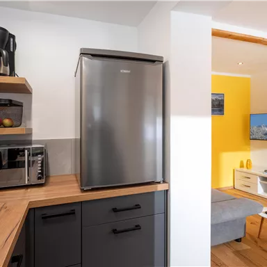 A modern kitchen with a refrigerator, a microwave, and a coffee machine. The room has a bright wood surface and an open transition to a living area with a television.