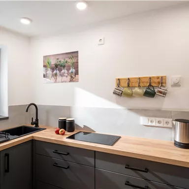 A modern kitchen with a wooden countertop and black furniture. A window lets in plenty of light and there are some kitchen utensils on the wall.