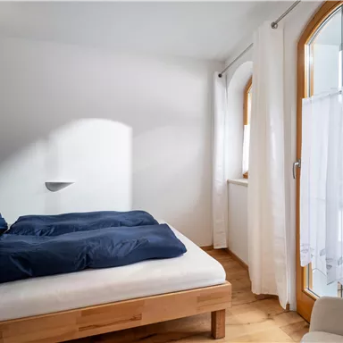 A cozy bedroom with a bed and blue pillows. Large windows provide plenty of light and a view outside.