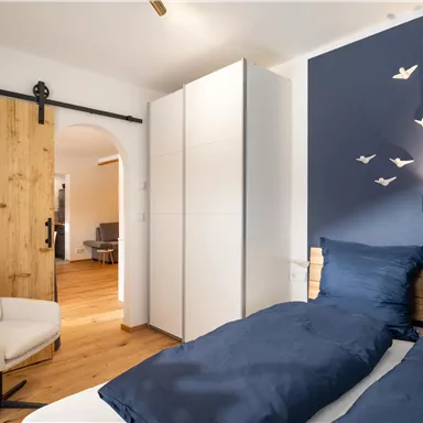 A modern bedroom with a blue accent wall and a white wardrobe. A comfortable chair is next to a bed with blue bedding.