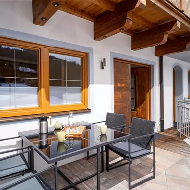 A cozy terrace with a table and chairs. In the background, large windows and a wooden door are visible.