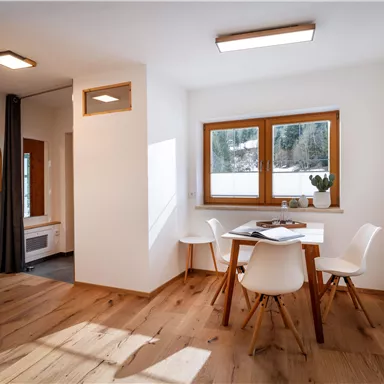 A modern dining room with a wooden table and white chairs. Large windows provide plenty of daylight and a view of nature.