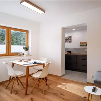 A cozy living space with a wooden table and modern chairs. The kitchen is visible in the background, and large windows let in a lot of light.