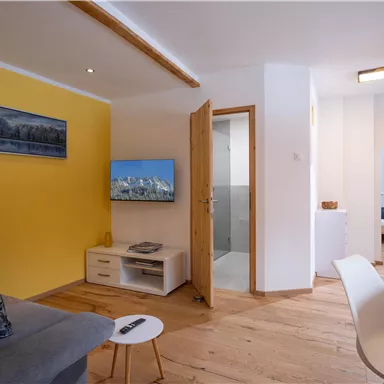 A modern living room with a gray sofa, a television, and a yellow wall. In the background, a bedroom and a bathroom can be seen.