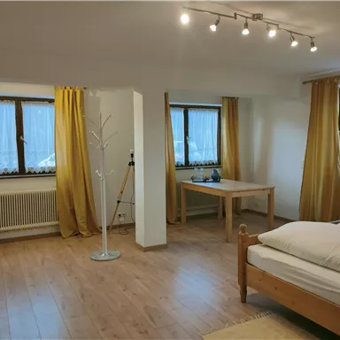 A bright room with wooden floors and large windows. The walls are neutral, and there is a table as well as a bed with bedding.