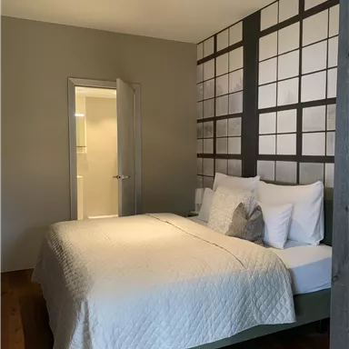 A modern bedroom with a large bed and neutral colors. The wall is designed with a graphic pattern, and a door leads to another room.