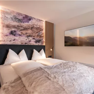 Ein modernes Schlafzimmer mit einem gemütlichen Bett und stilvollen Dekorelementen. An der Wand hängt ein Fernseher und ein künstlerisches Wandbild sorgt für eine angenehme Atmosphäre.
