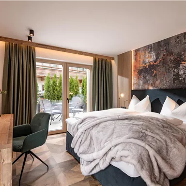 Ein modernes Schlafzimmer mit einem gemütlichen Bett und weichen Kissen. Große Fenster geben Blick auf eine grüne Terrasse und sorgen für helles Licht im Raum.