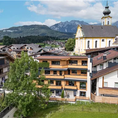 Eine malerische alpine Landschaft mit modernen und traditionellen Gebäuden. Im Hintergrund sind Berge und eine Kirche mit einer charakteristischen Kuppel zu sehen.