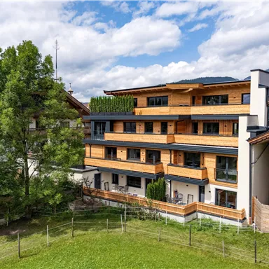 Ein modernes Gebäude mit Holzverkleidung und mehreren Balkonen. Im Vordergrund ist ein grüner Rasen mit einigen Bäumen zu sehen.