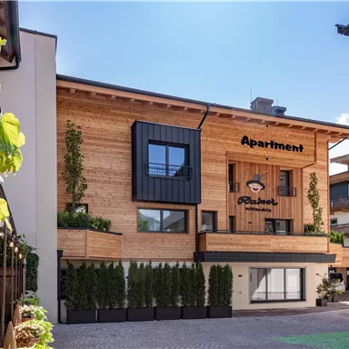 Ein modernes Appartementhaus mit Holzfassade und großen Fenstern. Der Eingang ist freundlich gestaltet, umgeben von grünen Pflanzen.