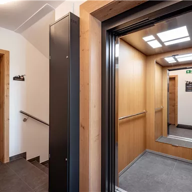 Ein moderner Aufzug in einem hellen Flur mit Holzverkleidung. Neben dem Aufzug befindet sich eine Treppe und eine Eingangstür.