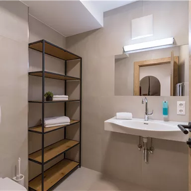 A modern bathroom with a bright atmosphere. It features a sink, shelves, and a shower.