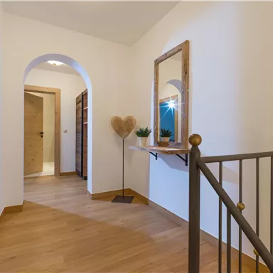 A bright hallway with a wooden floor and an elegant staircase. A mirror hangs on the wall and a heart-shaped decoration sits on a table.