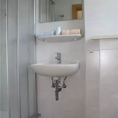 Ein modernes Badezimmer mit einer Dusche und einem Waschbecken. Auf dem Regal liegen Handtücher und der Boden ist mit einem orangen Badteppich bedeckt.