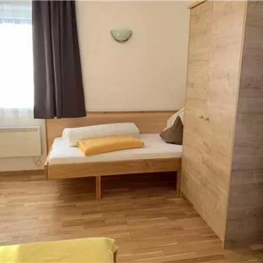 Ein einfaches Schlafzimmer mit einem Bett und einem Kleiderschrank aus Holz. Das Zimmer hat große Fenster und eine helle, freundliche Atmosphäre.