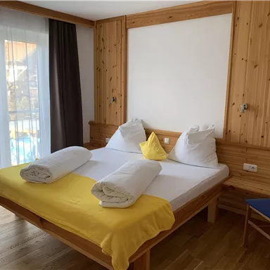 Ein gemütliches Schlafzimmer mit einem großen Bett, weißen Kissen und gelben Decken. Große Fenster lassen viel Licht herein und bieten einen Blick nach draußen.