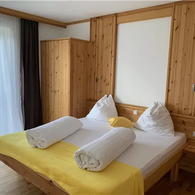 Ein gemütliches Schlafzimmer mit einem großen Bett und hölzernen Möbeln. Die gelbe Decke und die großen Fenster sorgen für eine einladende Atmosphäre.