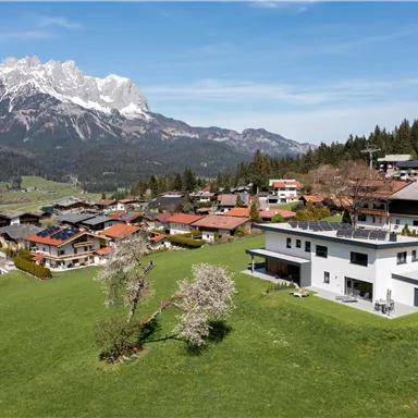 Eine malerische Berglandschaft mit schneebedeckten Gipfeln im Hintergrund. Im Vordergrund stehen moderne Häuser und eine grüne Wiese.