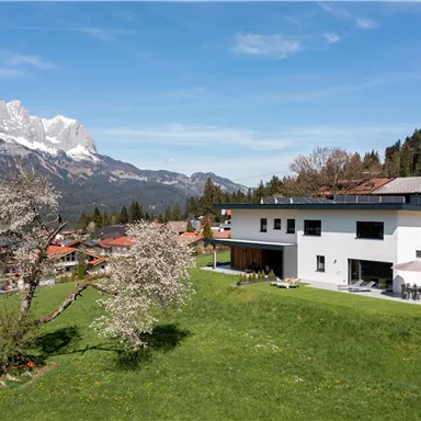 Ein modernes Haus in einer grünen Landschaft mit Blick auf die Berge. Die Umgebung ist malerisch mit Bäumen und kleinen Dörfern in der Nähe.