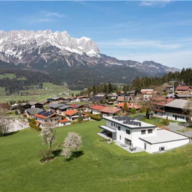 Eine malerische Landschaft mit schneebedeckten Bergen und einem ruhigen Dorf. Die Häuser liegen umgeben von grünen Wiesen und Bäumen.