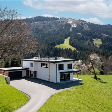 Ein modernes Haus auf einer grünen Wiese, umgeben von sanften Hügeln. Im Hintergrund erheben sich Waldgebiete und Berge unter einem klaren Himmel.