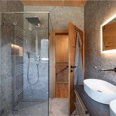 Ein modernes Badezimmer mit einer Glasdusche und Waschbecken. Die Wände sind in neutralen Grautönen gehalten, und Holzdetails sorgen für eine warme Atmosphäre.