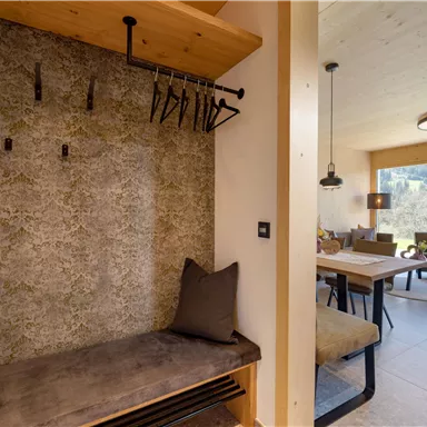 Ein moderner Eingangsbereich mit einer Sitzbank und Garderobenhaken. Im Hintergrund ist ein gemütlicher Wohnbereich mit Tisch und Stühlen sichtbar.