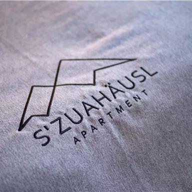 Ein modernes Logo für das Apartment "S'ZuaHäusl". Das Design ist schlicht und elegant auf einem grauen Hintergrund.